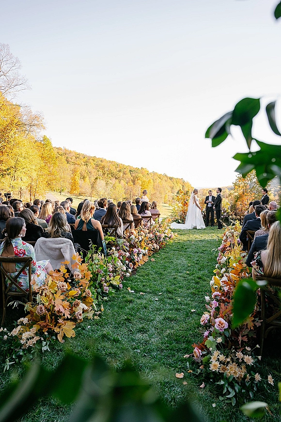 Wedding ceremony with aisle florals framing the bride and groom at a flowered arch on a grassy lawn with guests and fall trees under open sky