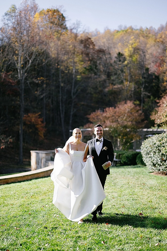 Couple portrait of bride and groom walking on a green lawn, bride in wedding dress and veil beside groom in tux, with fall trees and stone wall behind