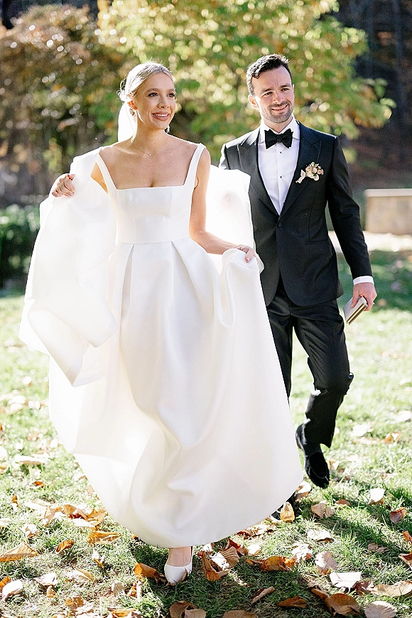 Couple portrait of bride and groom walking on a sunlit lawn with autumn trees, bride in strapless square neckline gown and veil, groom with vow booklet
