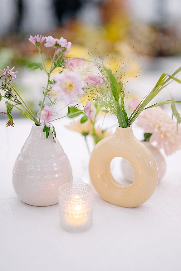 Wedding centerpieces with bud vase centerpiece in ceramic vases, pastel flowers and greenery beside a votive candle on a white tablecloth, bokeh lights behind