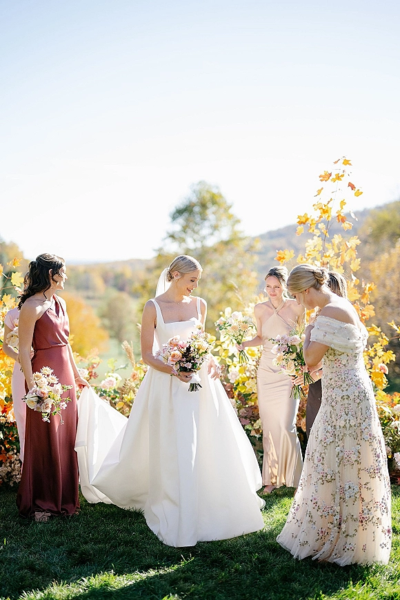 Bride with bridesmaids portrait as they stand on a grassy lawn, one adjusting her train, all holding bouquets with autumn hills behind them