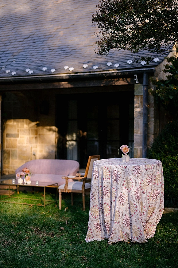 Cocktail table decor with patterned tablecloth and pink bud vase, beside lounge seating and votive candles on a lawn near a stone building