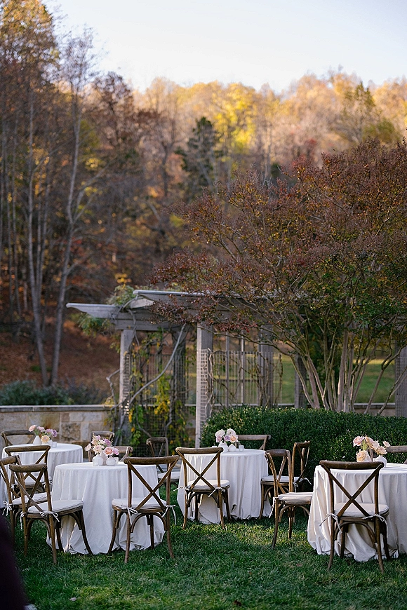 Outdoor reception tables with white linens on round wedding tables, cross back chairs, bud vases, and floral centerpieces on a fall lawn by a stone wall
