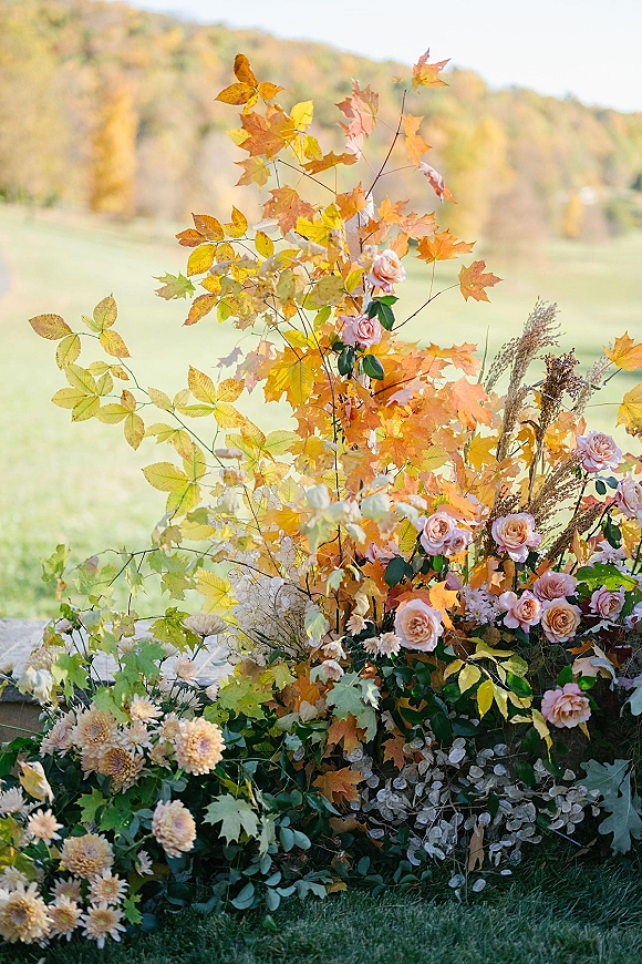 Wedding floral arrangement with fall wedding florals of blush roses, chrysanthemums, dried grasses, and branches on a lawn with rolling hills