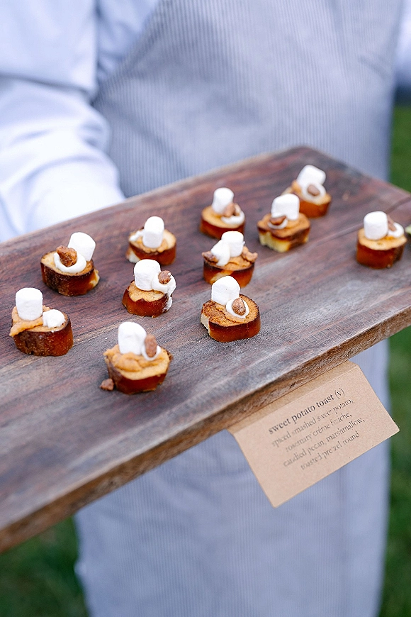Wedding passed appetizers on a wood serving board, passed hors d'oeuvres with marshmallow bites and a menu tag on an outdoor lawn