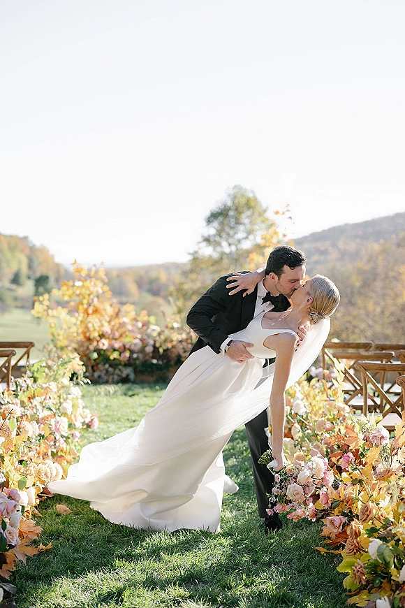 Wedding kiss portrait of groom dipping the bride in a veil and bouquet on a floral-lined aisle with wooden chairs and hills beyond
