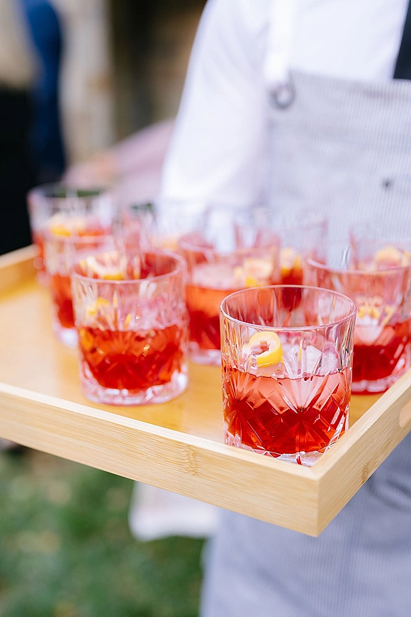 Wedding cocktails on a tray, red signature wedding cocktail with ice cubes and citrus peel garnish, carried by a server outdoors with greenery