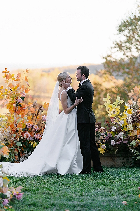 Couple portrait of bride and groom embrace, holding faces amid autumn garden florals, her cathedral veil flowing at sunset