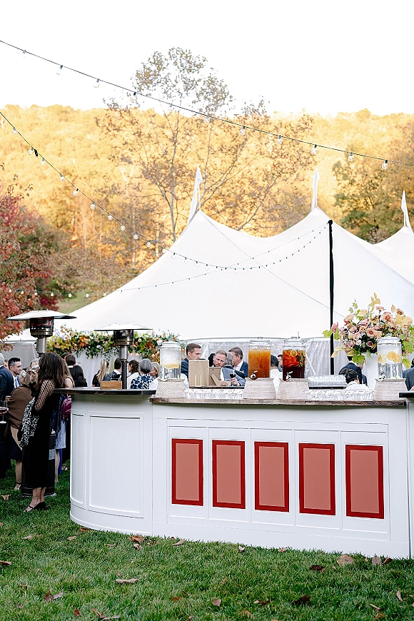 Wedding bar setup with iced drink dispensers and citrus garnish, floral accent and string lights under a white tent on a fall lawn