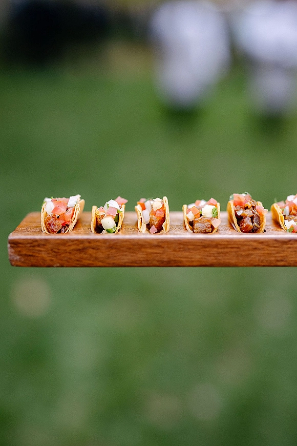 Wedding cocktail appetizers of mini tacos on a wooden serving tray with diced onion, tomato, and herbs, set on an outdoor lawn with blurred greenery