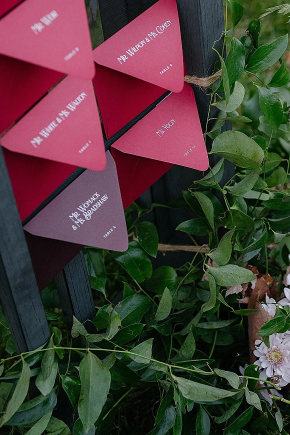 Wedding escort cards in red triangles with white text hang by twine on a black board, framed by greenery garland and flowers outdoors