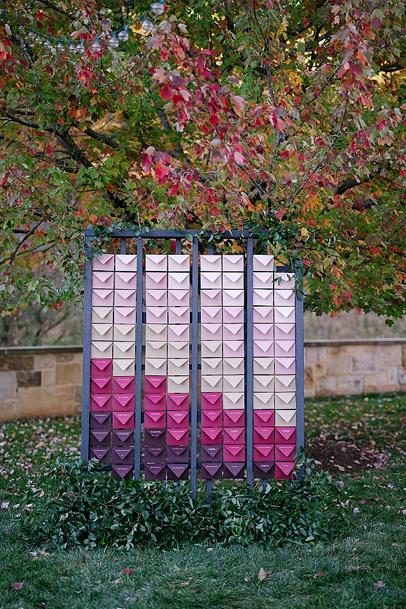 Wedding escort display with wedding escort card wall of pink ombre envelope escort cards on a stand with greenery garland, autumn trees behind