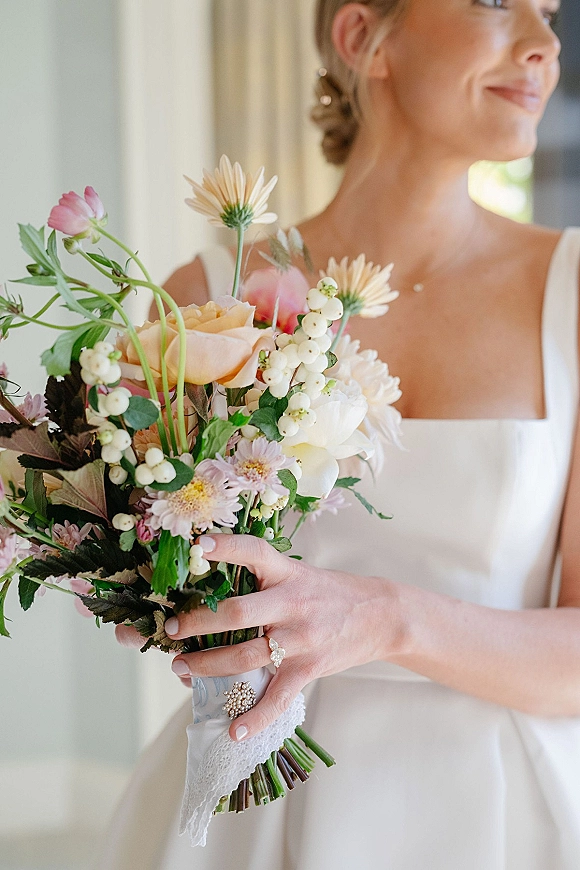 Bridal bouquet of pastel wedding flowers with roses, white berries and greenery, held in window light with lace ribbon and ring visible