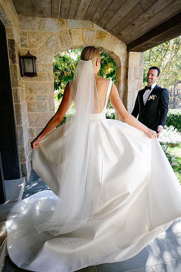 First look moment as bride approaches groom, holding her gown with cathedral veil and long train beneath a stone archway