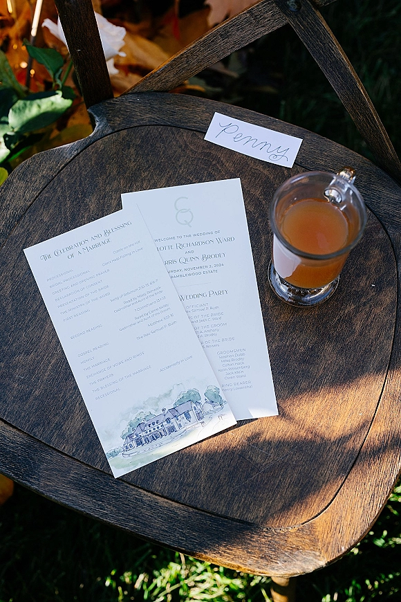 Wedding ceremony program and ceremony program design on a wooden chair with name card and glass mug, framed by greenery and florals outdoors