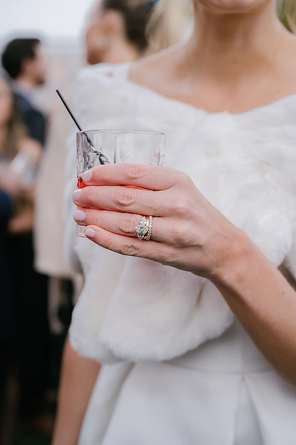 Wedding ring close-up of an oval diamond engagement ring and band stack on a bride’s hand holding a cocktail glass at an indoor reception