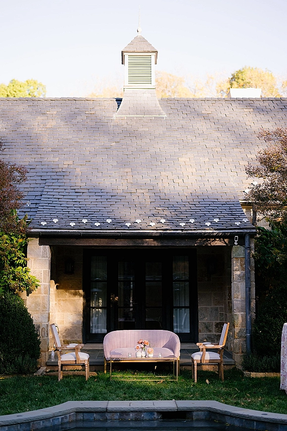 Lounge seating area wedding lounge setup with sofa, chairs, and a coffee table topped with floral centerpiece and candles on a patio by a stone house