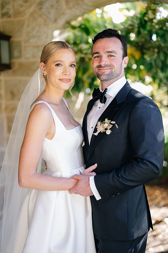 Couple portrait of bride and groom holding hands beneath a stone arch, her veil and earrings visible as greenery blurs behind them