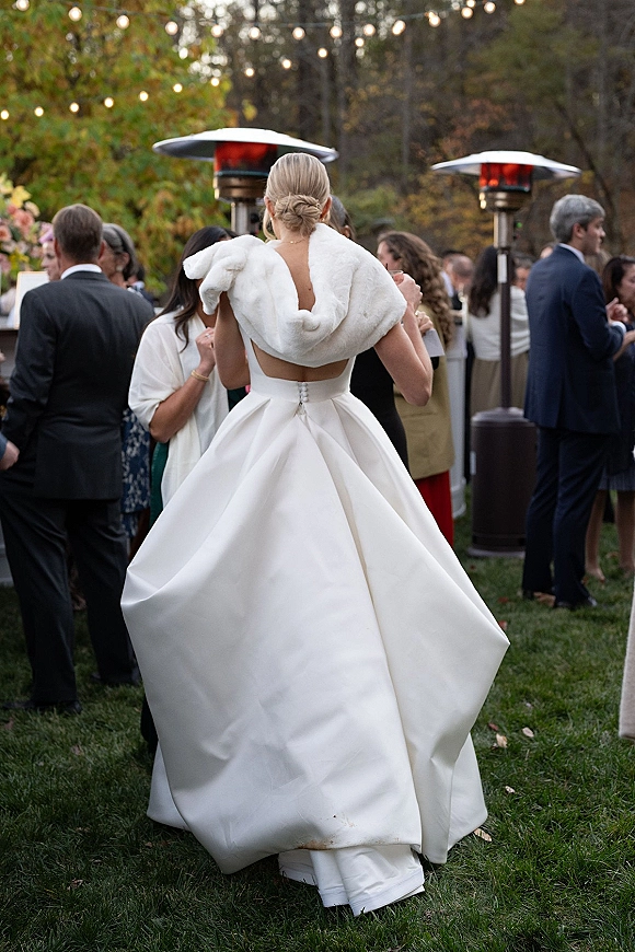 Bride reception moment in a strapless ball gown with fur stole and low bun, seen from behind under string lights on an evening lawn