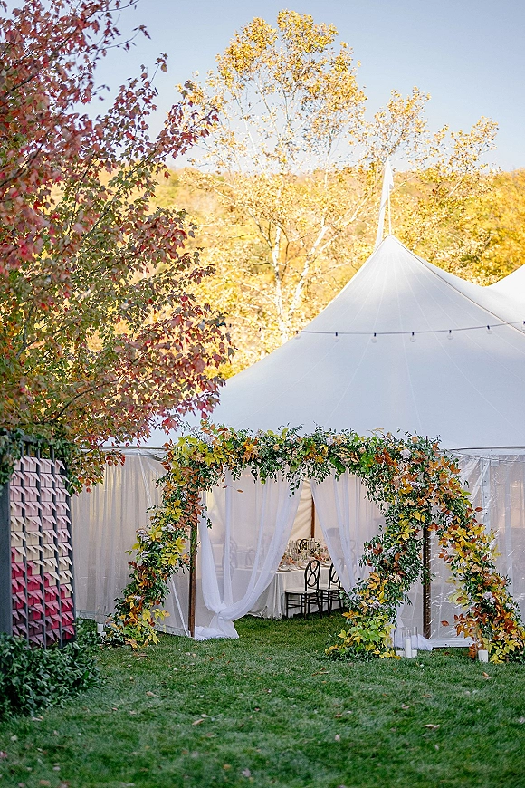 Wedding tent entrance with outdoor wedding tent draping, greenery garland, candles, and string lights under a clear tent amid autumn trees