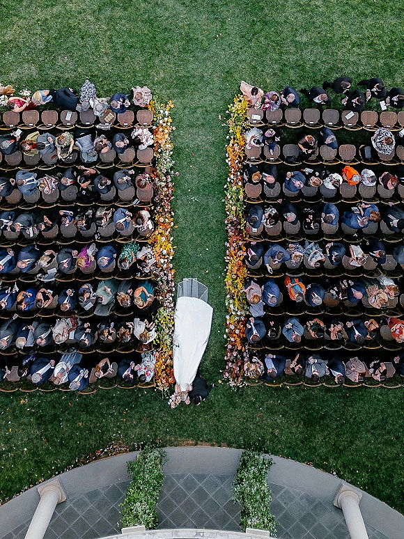 Ceremony aisle decor with fall foliage flowers lining an outdoor wedding aisle, as bride and groom stand amid chairs on lawn by columns