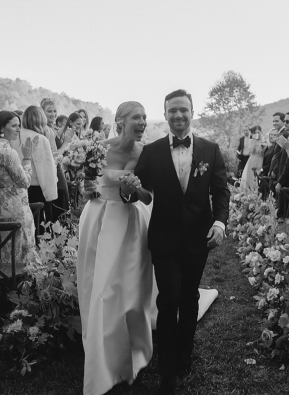 Wedding recessional as bride and groom walk the aisle holding hands, bouquet and veil flowing, with guests on a tree-lined lawn