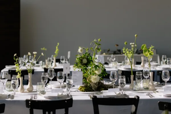 Reception tablescape with modern wedding table decor, styled with bud vases, against a sunlit white wall with black chairs.