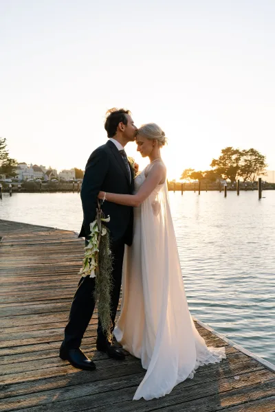 Couple portrait capturing a forehead kiss moment on a wooden dock at sunset, bride holding a white calla lily bouquet by the marina.