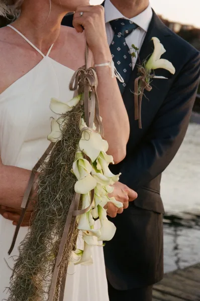 Bridal bouquet, a modern calla lily bouquet with long trailing ribbons, held on a dock beside tranquil water.