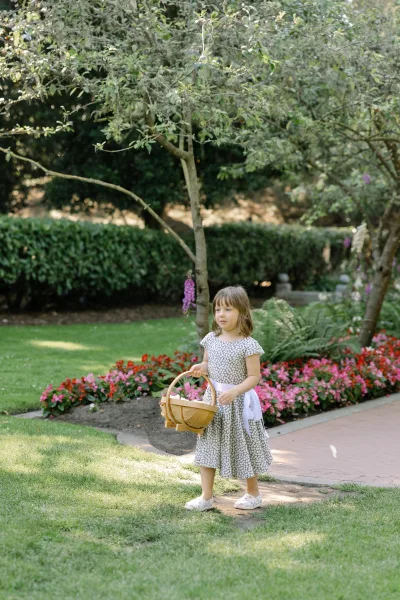 Flower girl with basket walks across a garden lawn and walkway, bordered by flower beds, trees, and hedges.