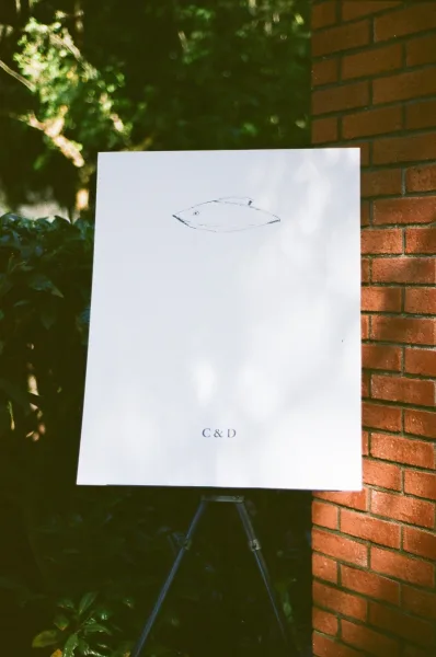 Wedding sign, a minimalist wedding sign featuring a bold monogram, set against a brick wall with surrounding greenery and shrubs.