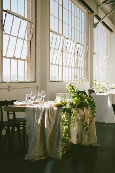 Sweetheart table decor with a cascading greenery table runner and glass hurricanes, framed by industrial windows and white walls.