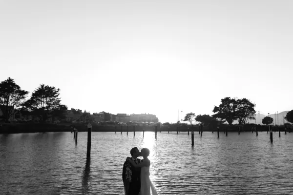 Couple portrait of a bride and groom embracing, the bride in a flowing bridal gown, by calm water at sunset with dock pilings and a city skyline.
