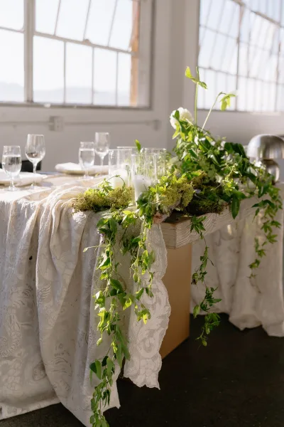 Reception tablescape featuring a greenery table runner and soft pillar candles, set in natural light by large windows against white walls.