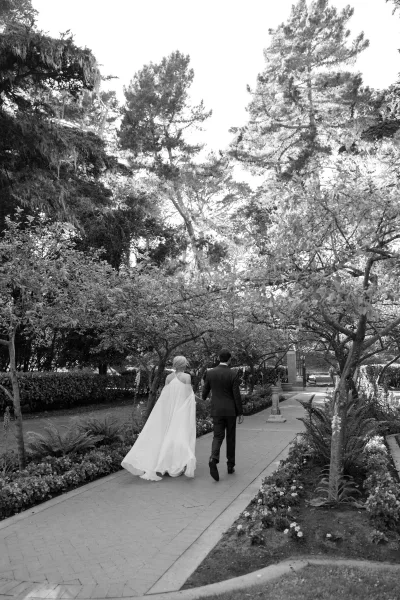 Couple portrait of the bride and groom walking along a tree-lined garden path, her simple flowing gown trailing as hedges and flowers border the way.