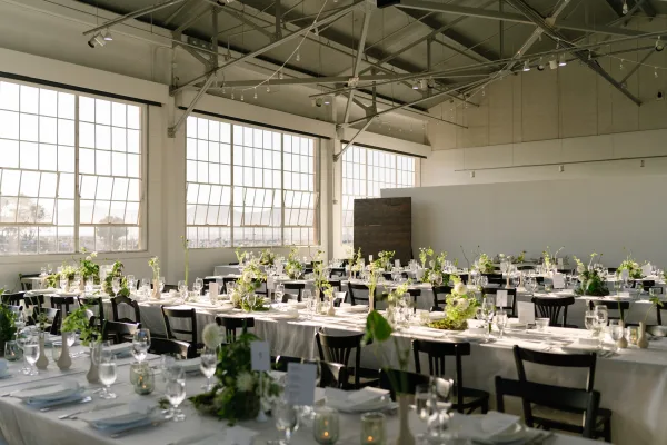 Reception tablescape on long banquet tables with minimalist centerpieces in bud vases, set beneath string lights and large industrial windows.