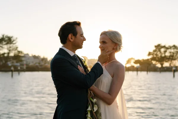 Couple portrait, an outdoor wedding portrait by the water at sunset, as the groom touches the bride's face and she holds a minimalist bridal bouquet.