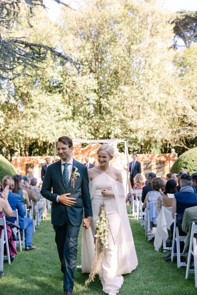 Ceremony recessional featuring a newlyweds aisle walk, hands clasped, bride in a modern bridal cape beside the groom on a garden lawn.