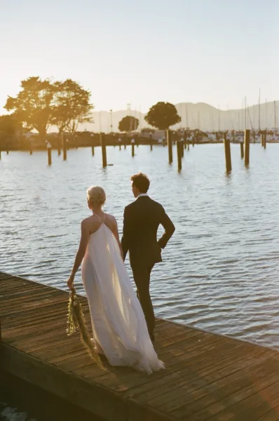 Couple portrait of a bride and groom walking along a wooden dock at sunset, her cape veil trailing as sailboat masts rise over the water.