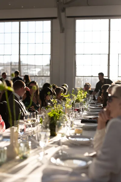 Reception tablescape on a long banquet table featuring floral centerpieces in an industrial loft framed by large windows.