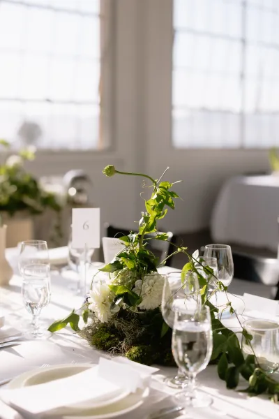 Reception tablescape with a greenery wedding centerpiece and white florals in a sunlit room with large windows and white walls.