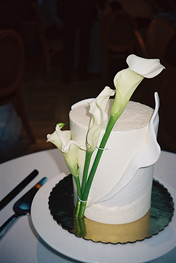 Wedding cake with a fondant drape topped with white calla lilies on a table, with cake knife and server against a dark wall