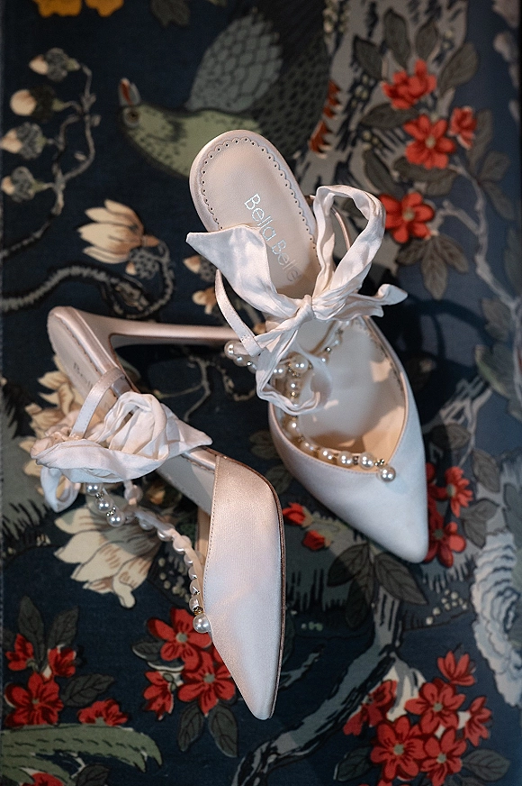 Bridal shoes, white satin heels with a satin bow and pearl details, arranged on floral patterned fabric over dark blue textile