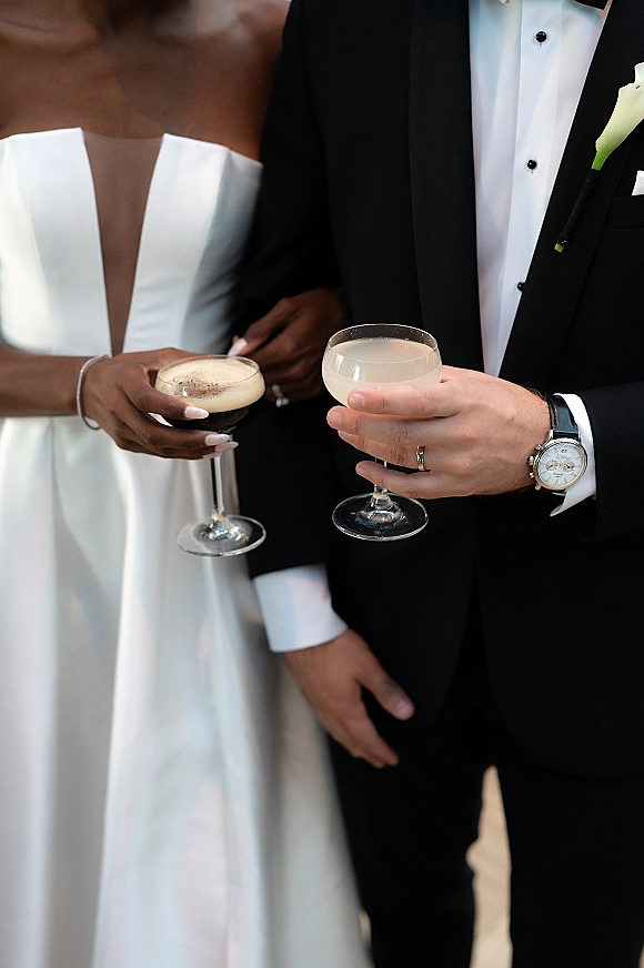 Wedding champagne toast as bride and groom clink champagne coupe glasses, showing satin dress, tuxedo, and rings in soft greenery light