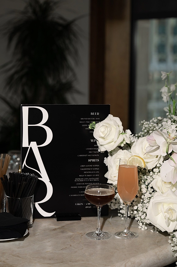 Wedding bar menu and signature cocktail menu on a black card beside white roses and orchids on a stone bar top by a window