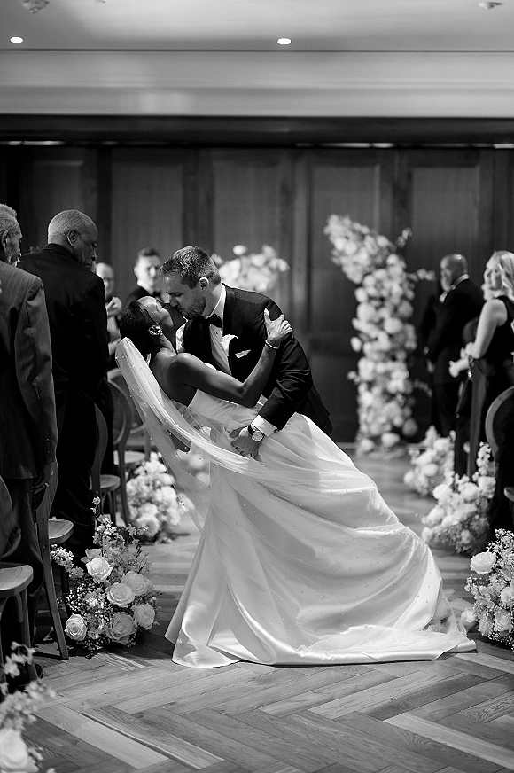 Wedding kiss as groom dips bride in strapless dress and cathedral veil on a floral-lined indoor aisle with guests and wood wall backdrop