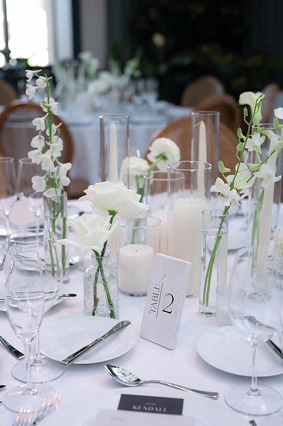Reception tablescape with a white wedding tablescape of roses in bud vases, glass candle cylinders, and place settings in an indoor venue