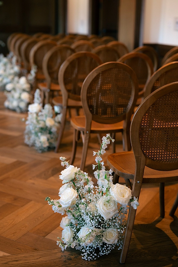 Ceremony aisle decor with wedding aisle flowers, white roses and baby's breath clusters along cane-back chairs on a herringbone wood floor