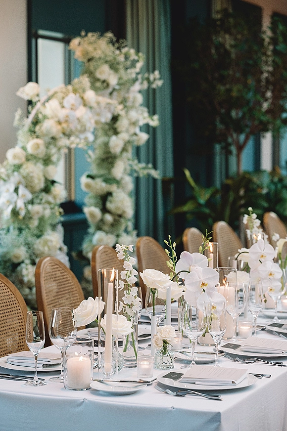 Reception tablescape wedding table setting with white orchid and rose centerpiece, taper candles, menus and glassware against dark paneled wall