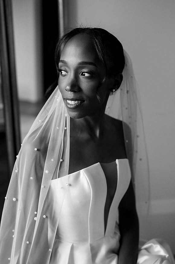 Bridal portrait in black and white of a bride by window light, looking away in a strapless gown and pearl-dotted veil near curtains